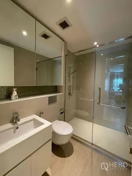 Condominium for rent in Watthana, Bangkok - Modern bathroom with mirrored cabinets, wall-mounted toilet, vanity sink, and glass-enclosed shower.