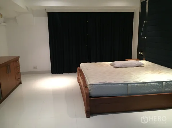 Condominium for rent in Khlong Toei, Bangkok - Large bedroom with wooden bed frame, white tile flooring and blackout curtains.