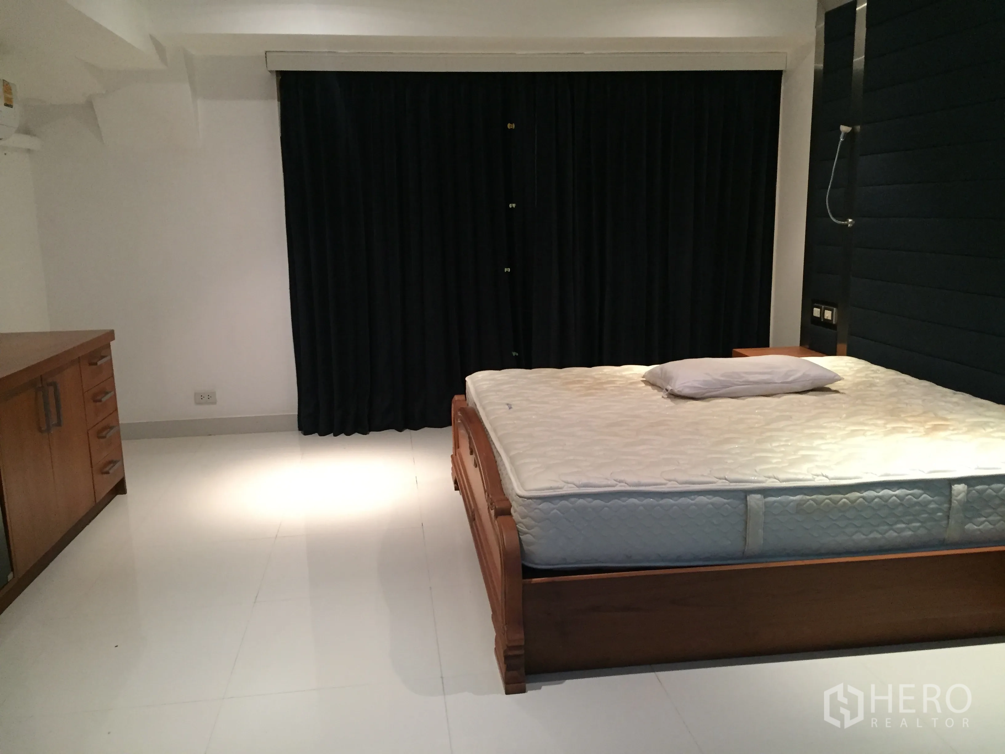 Condominium for rent in Khlong Toei, Bangkok - Large bedroom with wooden bed frame, white tile flooring and blackout curtains.