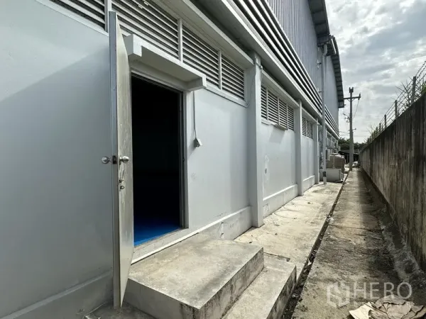 Factory for rent in Khlong Luang, Pathum Thani - Side service entrance of the factory with ventilation louvers and a concrete walkway along the boundary wall.