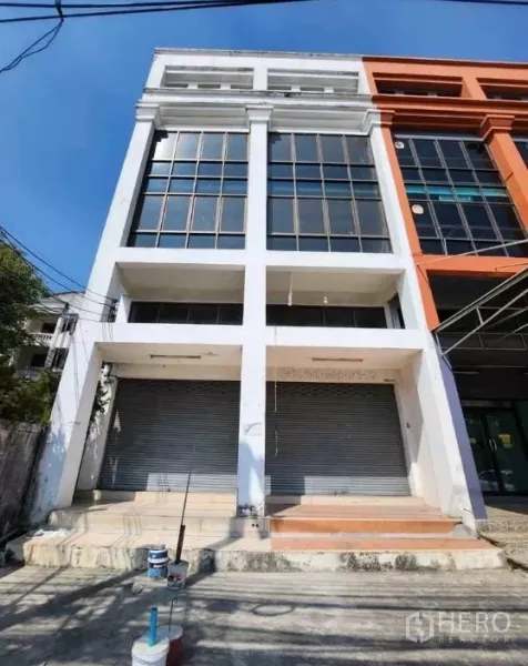 办公空间 出租在Suan Luang, Bangkok - 四层商业/办公建筑立面，大片落地窗与双卷闸门，蓝天背景。