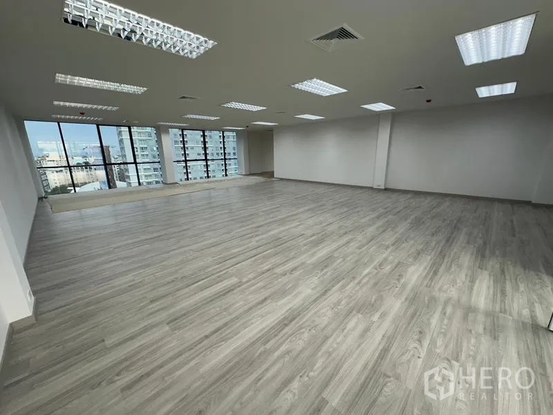 Office Space for rent - Spacious unfurnished office area with wood-look flooring, LED grid lighting, and a glazed facade overlooking nearby towers.