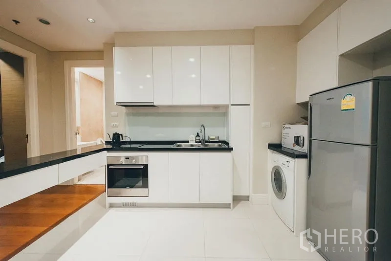 Condominium for rent in Khlong Toei, Bangkok - Contemporary white kitchen with black countertops, oven, fridge and washing machine.