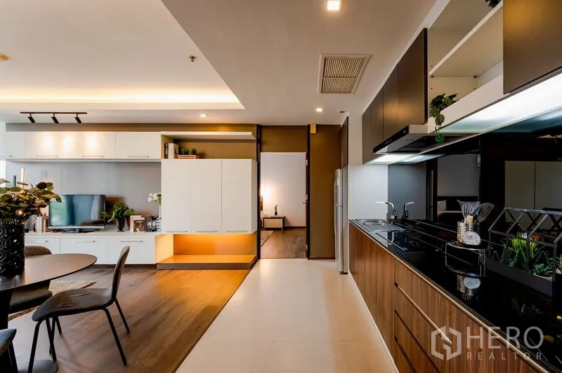 Condominium for sale in Khlong Toei, Bangkok - Open kitchen-to-living perspective showing storage cabinets, dining set, and doorway to a bedroom.