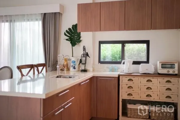Detached House for rent in Sai Mai, Bangkok - Modern L-shaped kitchen with white countertop, wood cabinetry and a window looking to greenery.