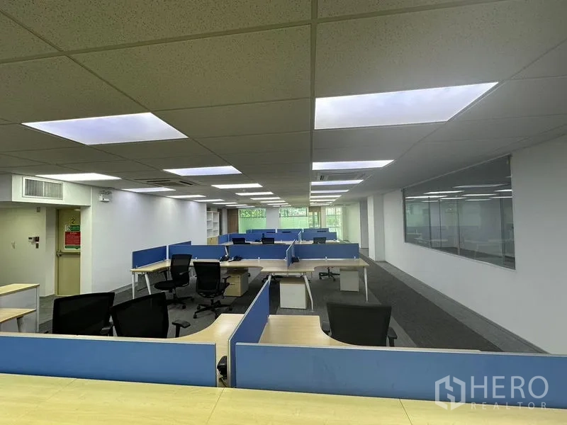 Office Space for rent - Rows of modular desks and swivel chairs under recessed lighting with a glass wall and greenery view ahead.