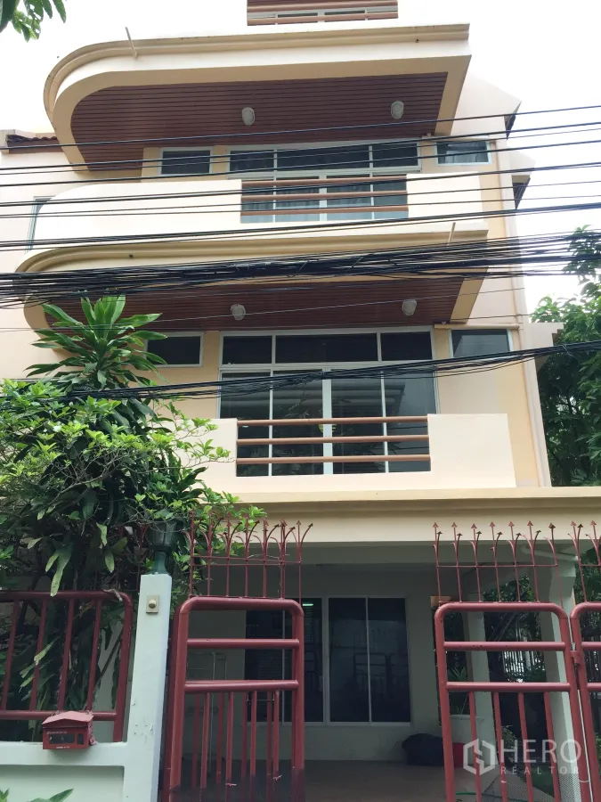 บ้าน ให้เช่าในWatthana, Bangkok - ด้านหน้าบ้านเดี่ยวสูงหลายชั้น มีระเบียงโค้ง กระจกบานใหญ่ และรั้วเหล็กสีแดง