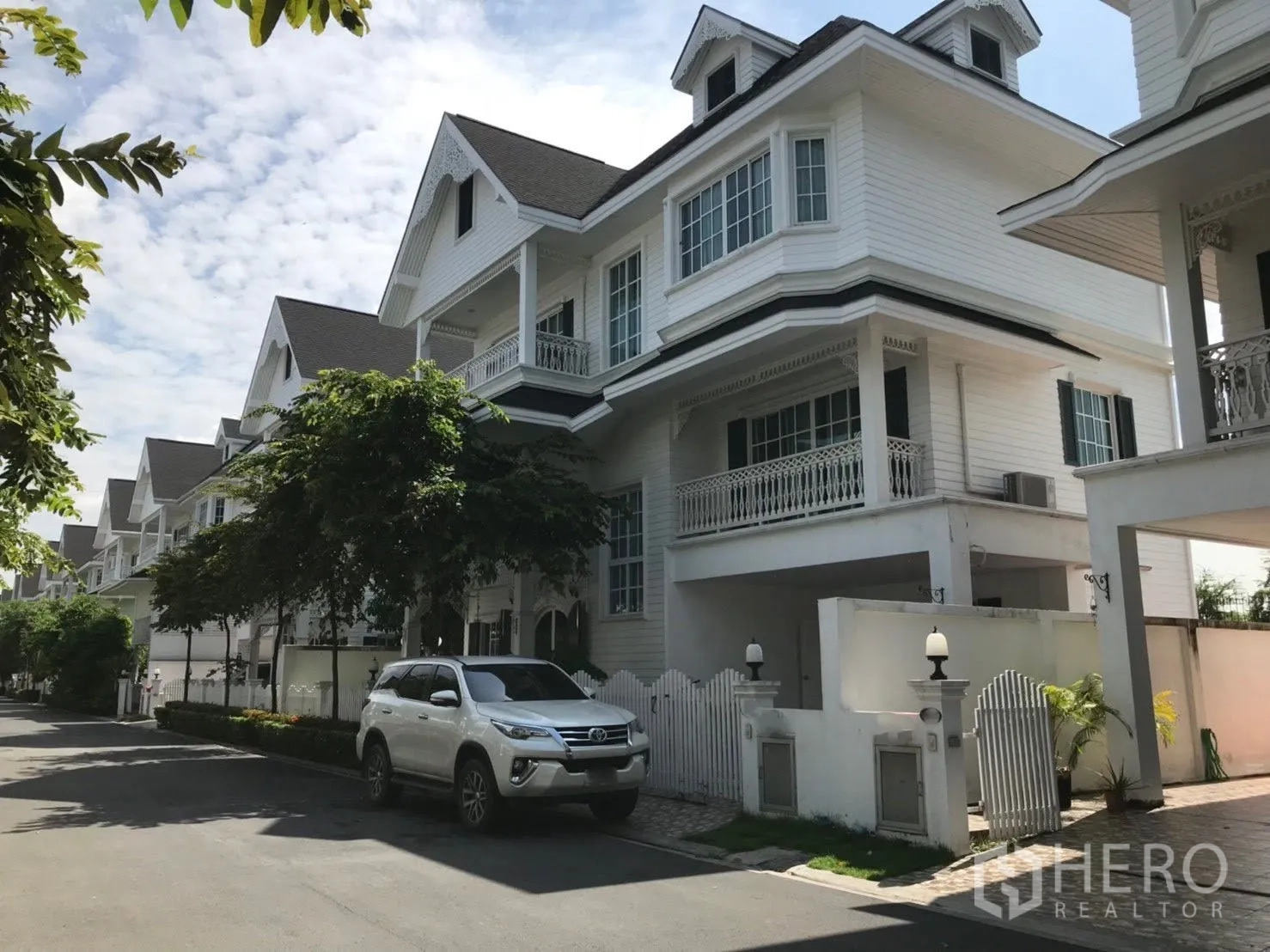 บ้านเดี่ยว for rent in Mueang Samut Prakan, Samut Prakan - แถวบ้านเดี่ยวสีขาวพร้อมระเบียงบนถนนในโครงการ มีรถเอสยูวีจอด