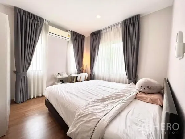 Detached House for rent or sale in Bang Khen, Bangkok - Corner bedroom with white bedding, study desk, air conditioner and double windows.