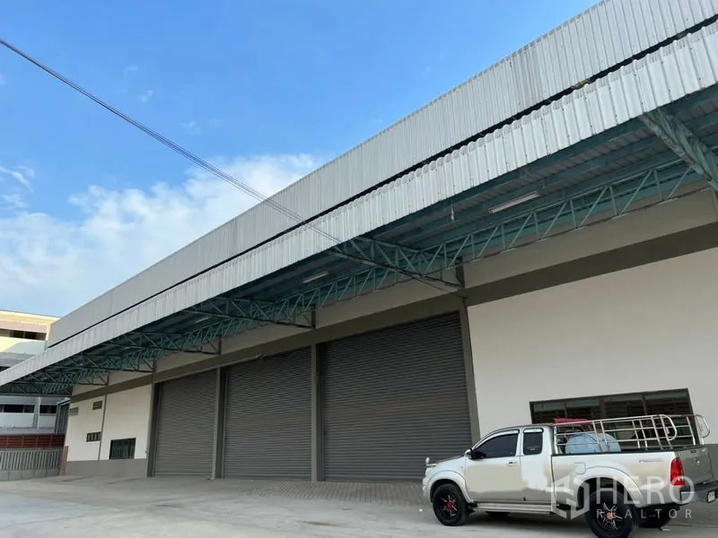 Warehouse for rent in Lat Krabang, Bangkok - Modern warehouse exterior with multiple roller shutter doors and a covered loading bay, pickup truck parked on the concrete yard.
