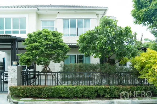 Detached House for rent or sale in Saphan Sung, Bangkok - Front facade with sliding windows, lush trees, trimmed hedges and a black metal fence.
