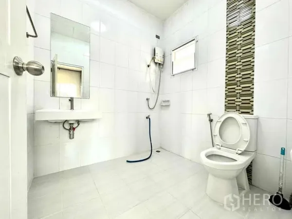 Detached House for rent in Lat Krabang, Bangkok - Clean white-tiled bathroom with shower water heater, toilet, sink and a small window.