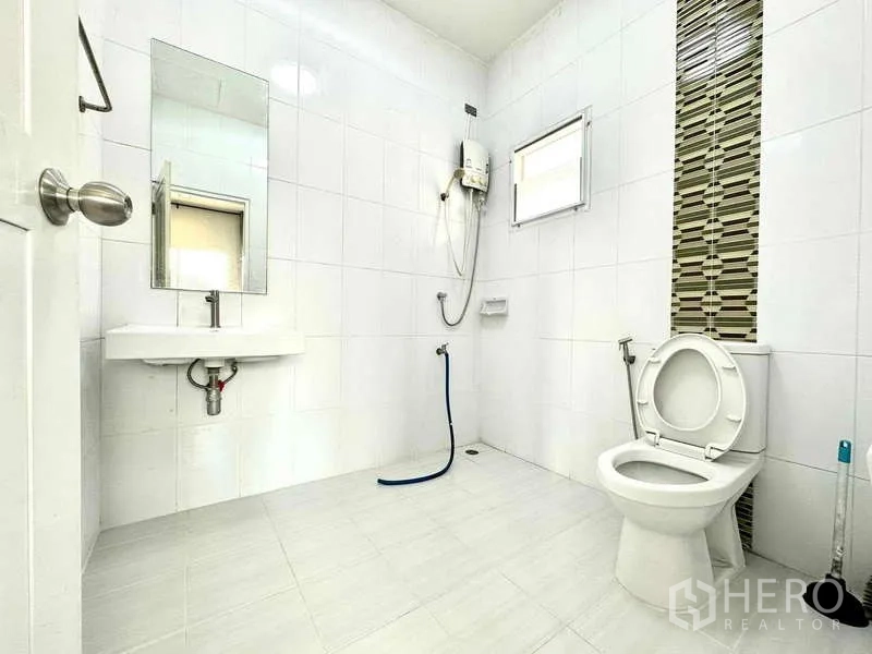 Detached House for rent in Lat Krabang, Bangkok - Clean white-tiled bathroom with shower water heater, toilet, sink and a small window.