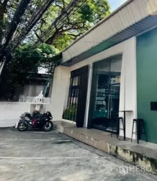 零售空间 出租在Watthana, Bangkok - 店铺侧面视角，有停车位与摩托车，玻璃门店面与大树