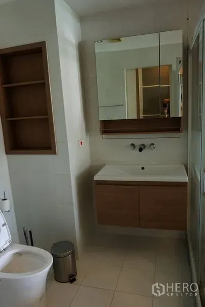 Condominium for sale in Khlong Toei, Bangkok - Modern bathroom with wall-mounted vanity, mirrored cabinet, built-in shelving and glass shower.