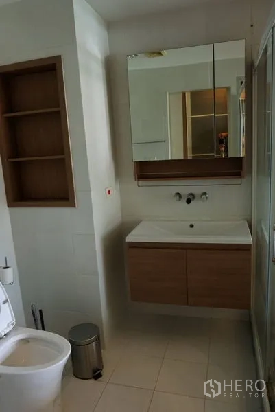 Condominium for sale in Khlong Toei, Bangkok - Modern bathroom with wall-mounted vanity, mirrored cabinet, built-in shelving and glass shower.