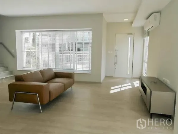 Townhouse for rent in Prawet, Bangkok - Open-plan lounge showing bay window, staircase and front door with natural light across wood-look flooring.