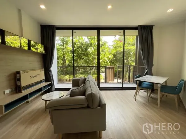 Condominium for sale in Watthana, Bangkok - Bright living room with gray sofa, dining set, and floor-to-ceiling windows opening to a leafy balcony view.