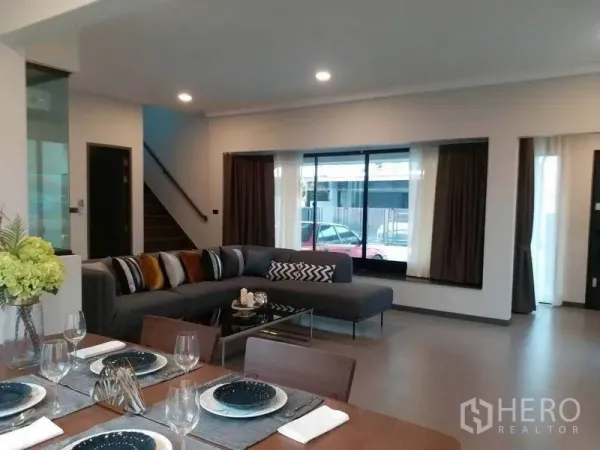 Townhouse for rent in Bang Phli, Samut Prakan - Spacious lounge facing a staircase, L-shaped sofa, and dining table by wide picture windows.
