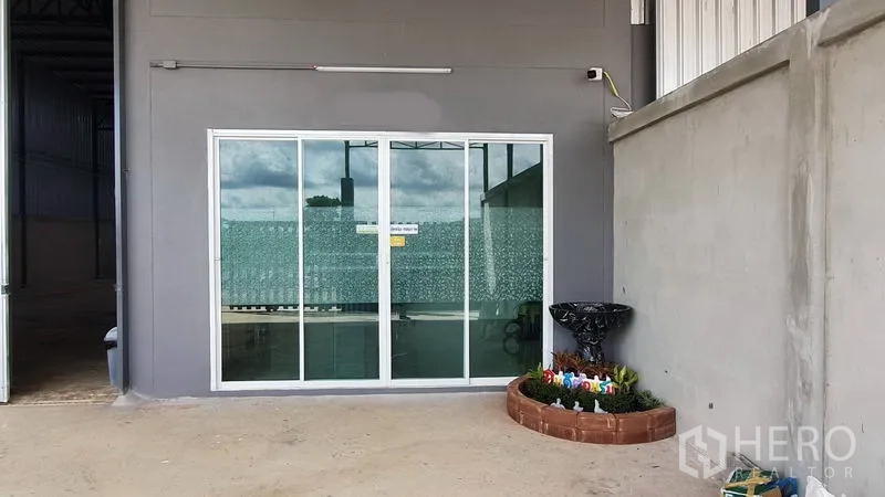 Warehouse for rent in Lam Luk Ka, Pathum Thani - Ground-level glass sliding door to office next to concrete yard and small planter.
