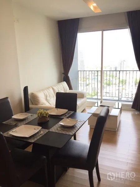 Condominium for rent in Khlong Toei, Bangkok - Dining area beside a bright living room with sofa and floor-to-ceiling window opening to a city-view balcony.