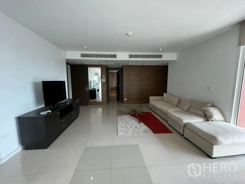 公寓 出租在Watthana, Bangkok - 起居室朝入口视角，米色转角沙发、电视柜与吊顶中央空调。