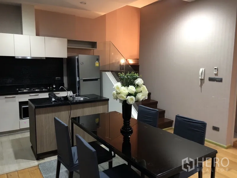 公寓 出租或出售在Khlong San, Bangkok - 现代厨房，黑色石材台面、白色柜体，旁边是玻璃扶手楼梯与餐桌。
