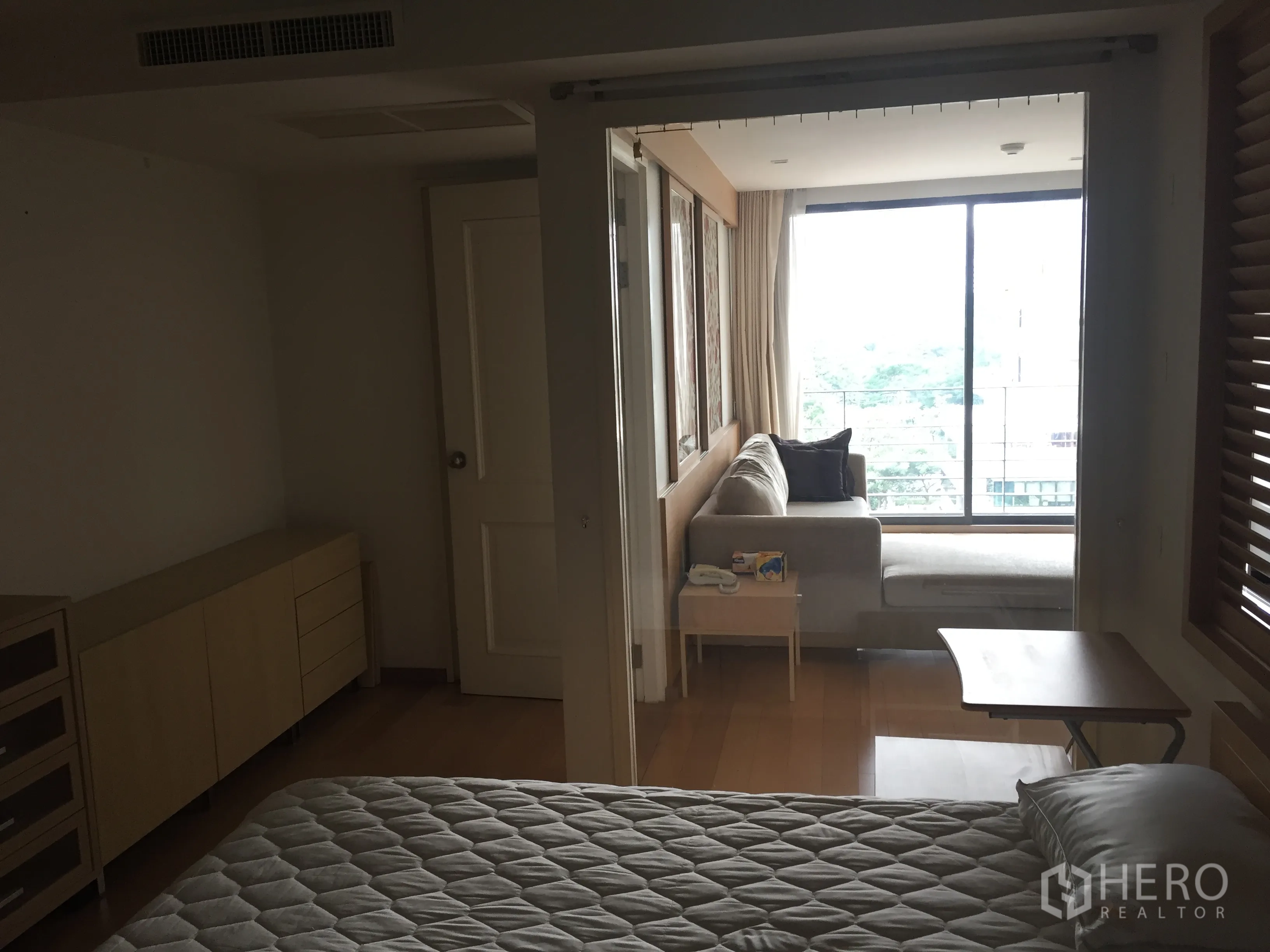 公寓 出租在Pathum Wan, Bangkok - 卧室通过推拉玻璃与客厅相连，转角沙发靠近落地窗，享绿色与城市景观。