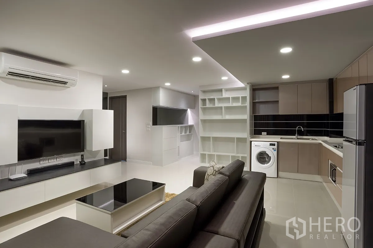 Condominium for rent in Khlong Toei, Bangkok - Living area showing TV wall, built-in shelving, and kitchen with fridge and washing machine.