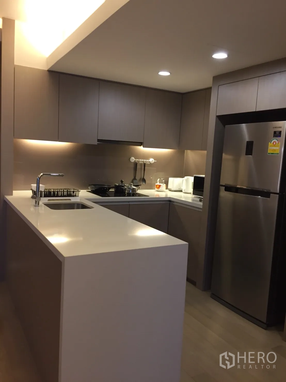 公寓 出租在Watthana, Bangkok - 现代U型厨房，白色中岛台面，嵌入式柜体与不锈钢冰箱。