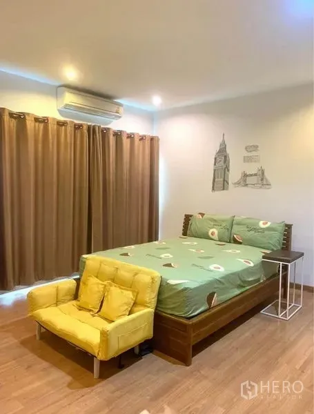 Townhouse for rent in Suan Luang, Bangkok - Primary bedroom with double bed, yellow sofa, wood flooring, air conditioner and blackout curtains.