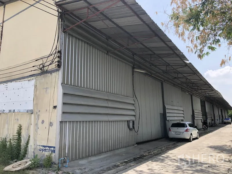 Warehouse for rent in Lam Luk Ka, Pathum Thani - Row of metal-clad warehouse units with large sliding doors, covered loading area and driveway with parked car in Lam Luk Ka.