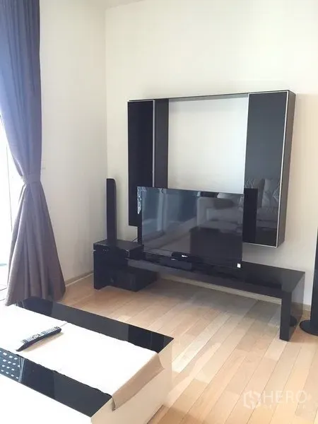 Condominium for rent in Khlong Toei, Bangkok - Modern living area with black media wall and flat-screen TV next to large curtained window.
