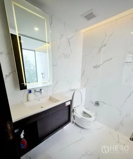 Condominium for rent in Khlong Toei, Bangkok - Sleek second bathroom with white marble walls, vanity and backlit mirror.