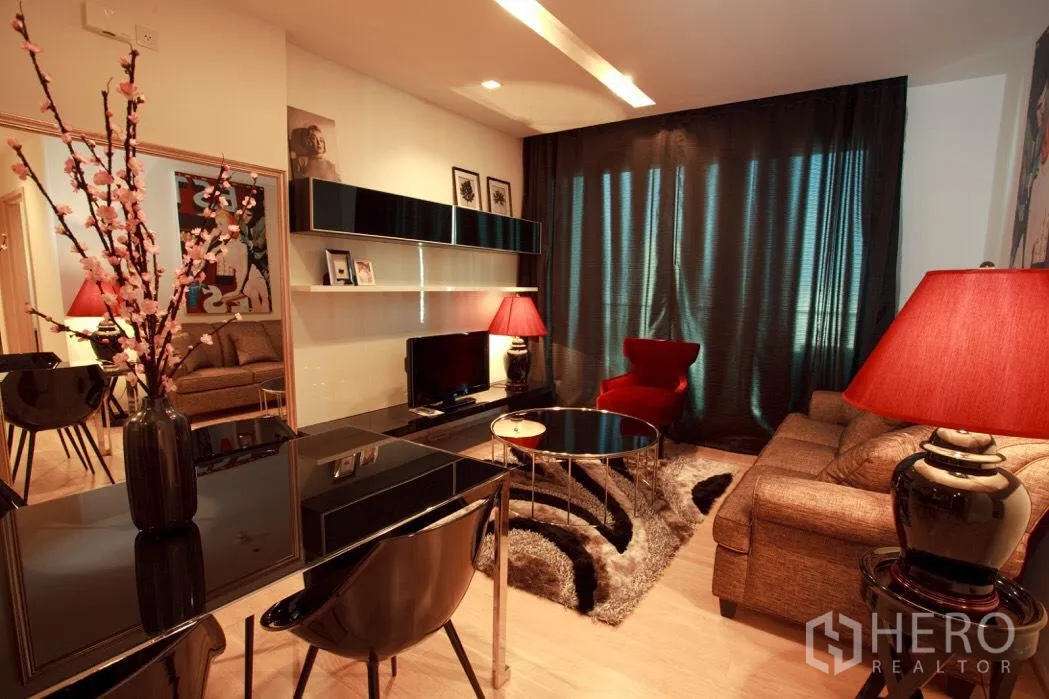 Condominium for rent in Khlong Toei, Bangkok - Warm living room with dark curtains, sectional sofa, red accent chair, TV console and floating shelves.
