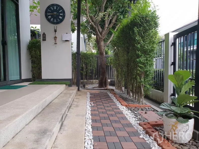 Detached House for rent in Bang Kapi, Bangkok - Side garden with paved walkway bordered by white pebbles, mature tree, hedges, and sliding glass door.