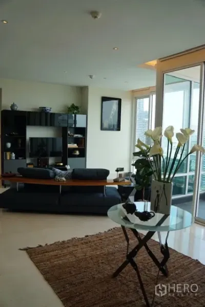 Condominium for rent in Bang Rak, Bangkok - Spacious living room with sofa set, TV wall, sliding doors to a balcony, and a glass accent table with flowers.