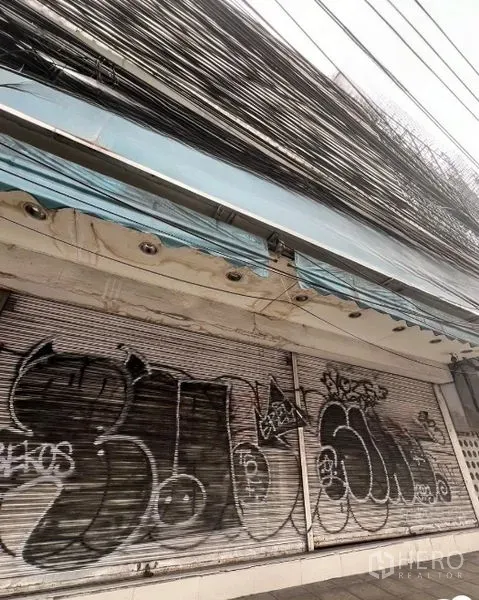 Shophouse for rent in Watthana, Bangkok - Wider exterior view of the shuttered storefront under a blue awning and dense utility lines.