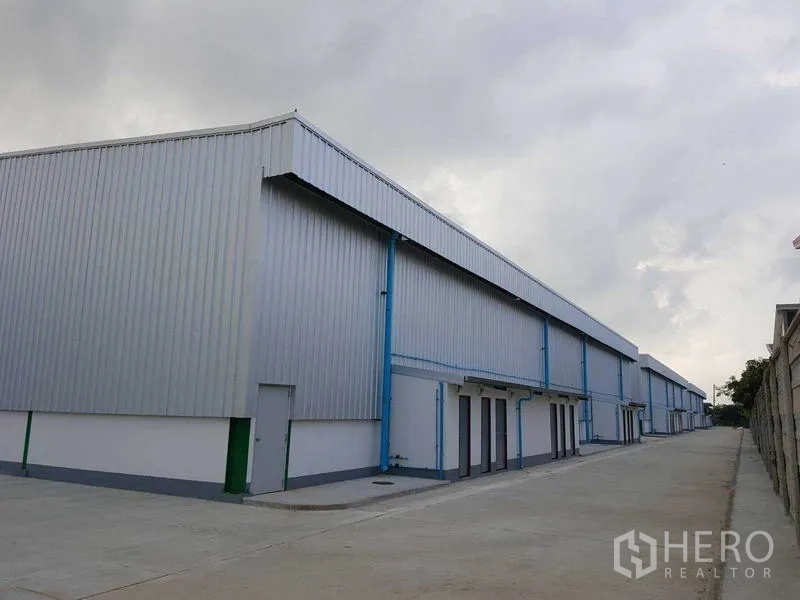 Warehouse for rent in Prawet, Bangkok - Side view of metal-clad warehouse buildings along a spacious concrete road with secure perimeter wall.