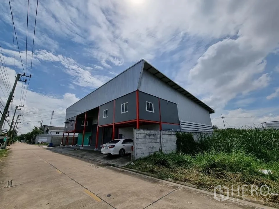 Warehouse for rent in Lam Luk Ka, Pathum Thani - Corner street view of a metal-clad warehouse along a wide access road and power lines.
