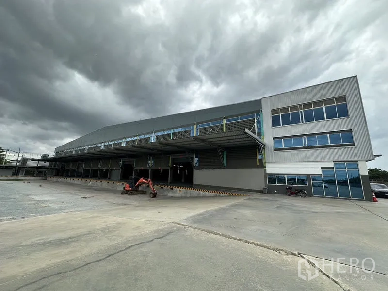 Factory for rent in Bang Sao Thong, Samut Prakan - Modern warehouse and office facade with covered dock loading bays and a wide truck yard under cloudy skies.