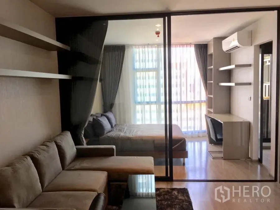 Condominium for rent in Watthana, Bangkok - Open-plan living room and bedroom divided by a glass wall with a grey sofa and floor-to-ceiling windows.