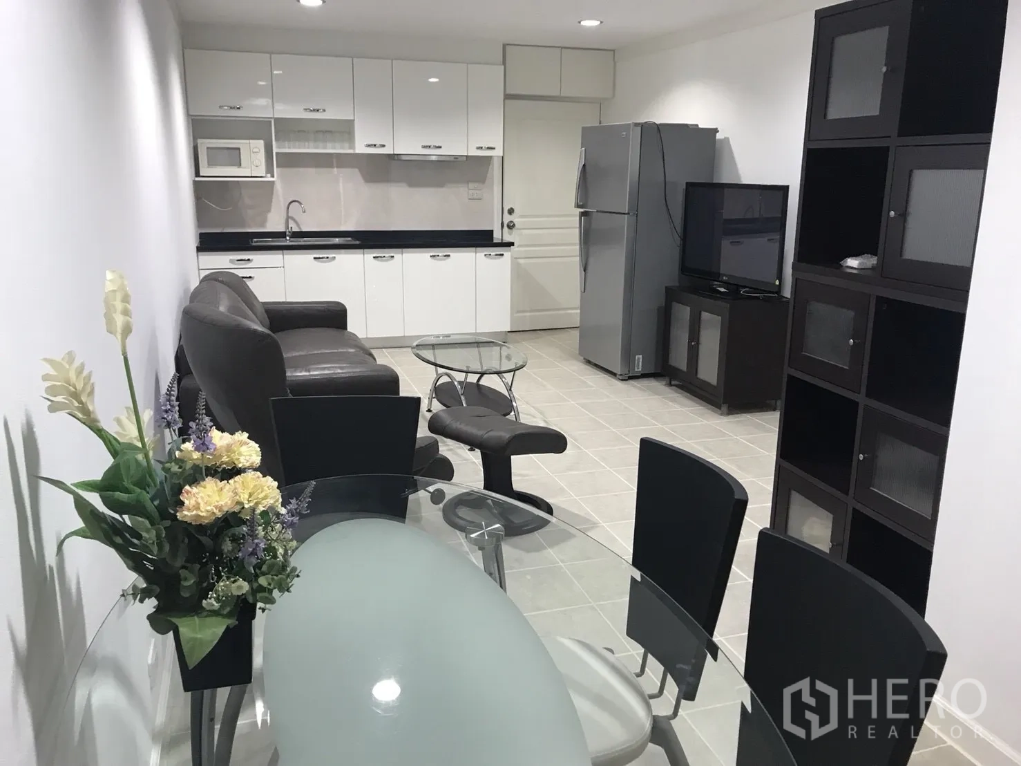 公寓 出租在Khlong Toei, Bangkok - 开放式客厅与白色小厨房，配玻璃餐桌、皮沙发、电视柜与大冰箱
