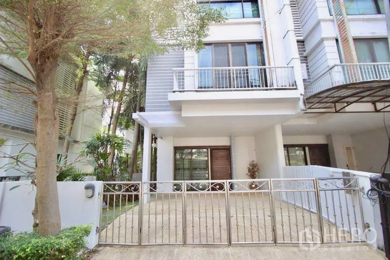Townhouse for sale in Suan Luang, Bangkok - Modern three-storey townhouse facade with gated driveway and balcony, shaded by trees.