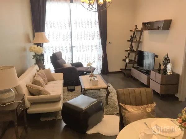 Condominium for rent in Watthana, Bangkok - Warm living room with sofa, TV unit, shelves, dark wood floors, and sheer curtains over tall windows.