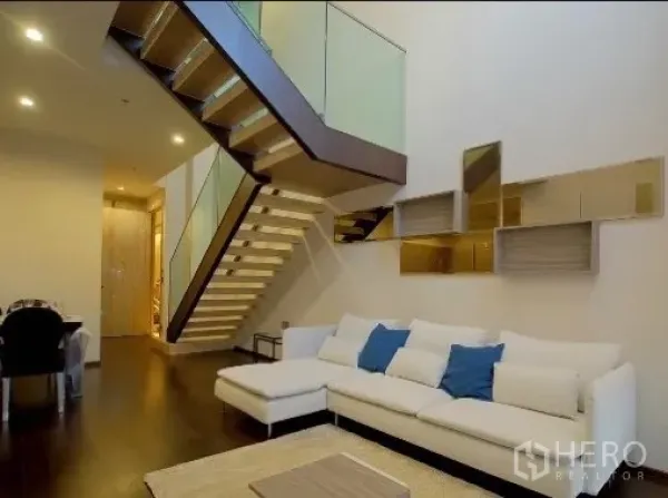 Condominium for rent in Watthana, Bangkok - Duplex living room with double-height ceiling, white L-shaped sofa and glass-balustrade staircase.