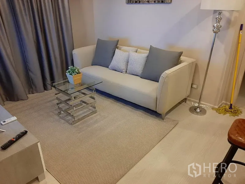 Condominium for rent or sale in Khlong Toei, Bangkok - Living room with beige sofa, rug, glass coffee table and tall floor lamp.