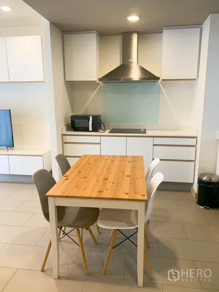 Condominium for rent in Watthana, Bangkok - Open-plan kitchen with white cabinetry, range hood, cooktop, microwave and a wooden dining table for four.