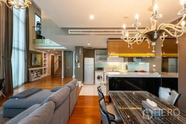 Condominium for rent in Khlong Toei, Bangkok - Double-height living and dining area with gray sectional, chandeliers, and an open kitchen.