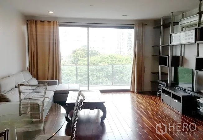 Condominium for rent in Khlong Toei, Bangkok - Open-plan living room with floor-to-ceiling glass doors to a balcony, built-in TV wall, and sofa set overlooking trees.